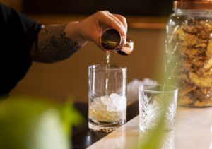 A bartender pours a measured amount of whiskey from a jigger into a large mixing glass with ice. There is a green plant in the foreground.