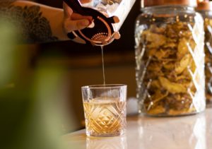 A bartender pours whiskey from a copper strainer into an etched glass with one large whiskey cube in the glass. The glass sits atop a white marble bar and there is a green plant in the foreground.