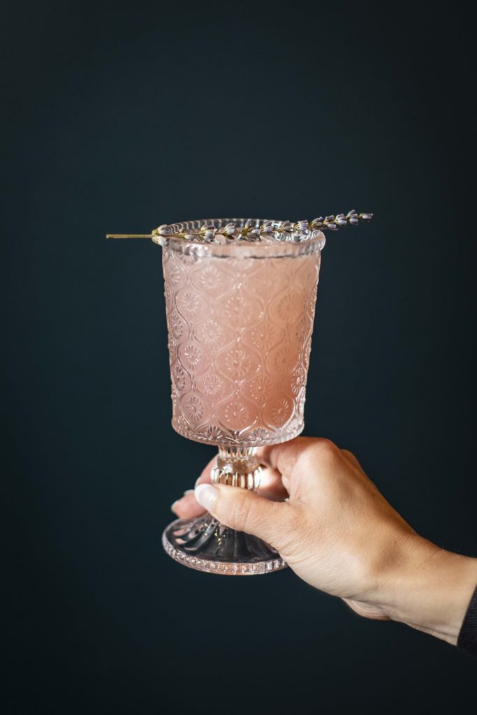 A woman’s hand is holding a blush pink drink in an etched tall glass. The drink is being held in front of a dark blue wall. There is a sprig of lavender atop the glass.
