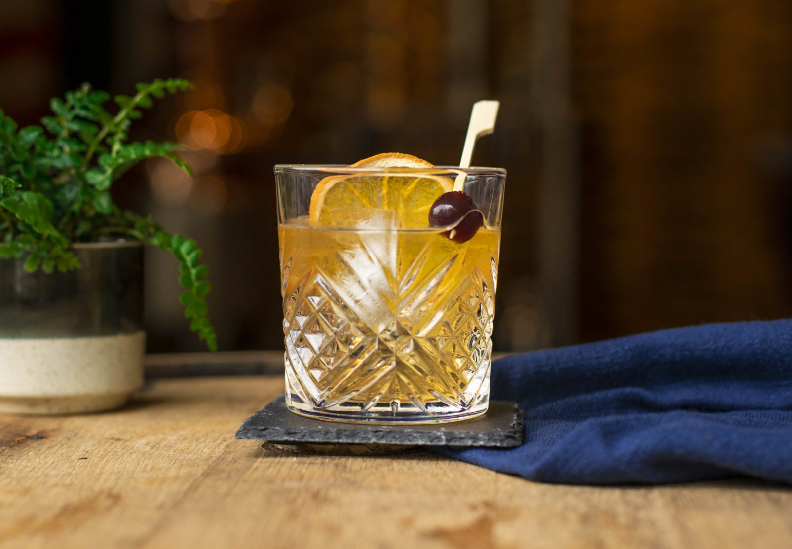 An Old Fashioned whiskey cocktail sits atop a wooden barrel with a stone coaster underneath and a royal blue napkin next to the drink. The cocktail has one large ice cube in it, as well as a slice of dried orange, and a bourbon cherry on a swizzle stick