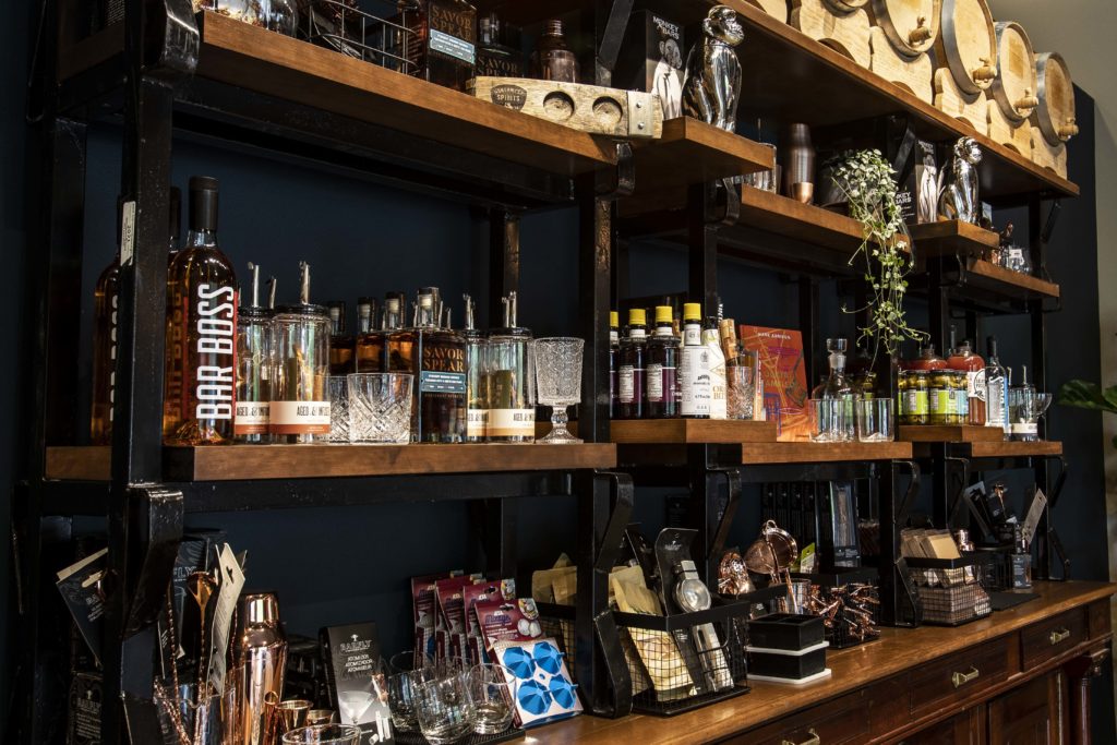 A well-stocked merchandise shelf is filled with various tools, books, and trinkets for the at-home bar enthusiast