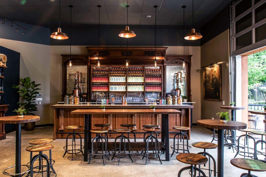 Image of an inviting tasting room. There is an impressive selection of liquor bottles lined up neatly on a Mahogany wooden display behind a white bar top. There are a few wooden tables with wooden stools. There is an industrial, yet warm feeling in the space.