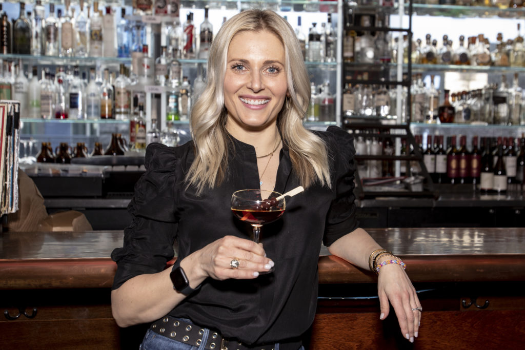 Portrait of Missy Firnstahl-Claridge, owner of Northwest Spirits. She is holding a Manhattan cocktail in front of a bar lined with hundreds of liquor bottles