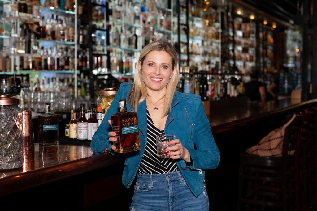 Portrait of Missy Firnstahl-Claridge, owner of Northwest Spirits. She is holding a bottle of Savor Spear Straight Bourbon Whiskey in front of a bar lined with hundreds of liquor bottles