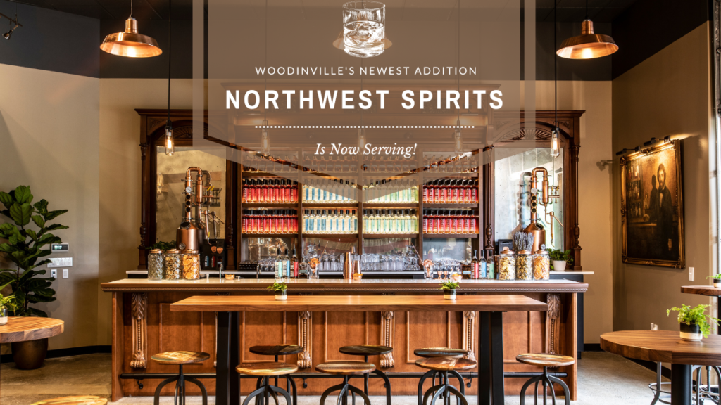 Image of an inviting tasting room. There is an impressive selection of liquor bottles lined up neatly on a Mahogany wooden display behind a white bar top. There are a few wooden tables with wooden stools. There is an industrial, yet warm feeling in the space. A text overlay reads "Woodinville's Newest Addition Northwest Spirits is now serving!"