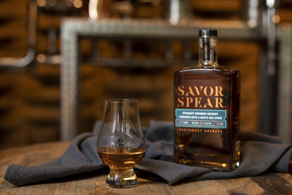 Image of a glass of bourbon sitting next to the bottle it was poured from, Savor Spear Straight Bourbon Whiskey. The glass and bottle sit atop a wooden barrel in front of a liquor still.