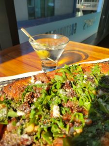 Image of a delicious looking fresh pizza topped with bright green arugula, fig, and prosciutto. There is a gin martini with an olive beside it.