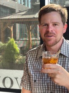 Our Master Distiller, Nick Warren, sits with a whiskey in hand in front of a window. There is a smirk on his face