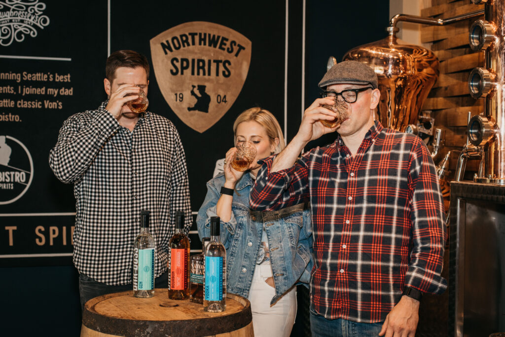 Three people are standing together sipping a glass of whiskey. In front of them is a barrel with 3 liquor bottles. Behind them is a sign that reads "Northwest Spirits"