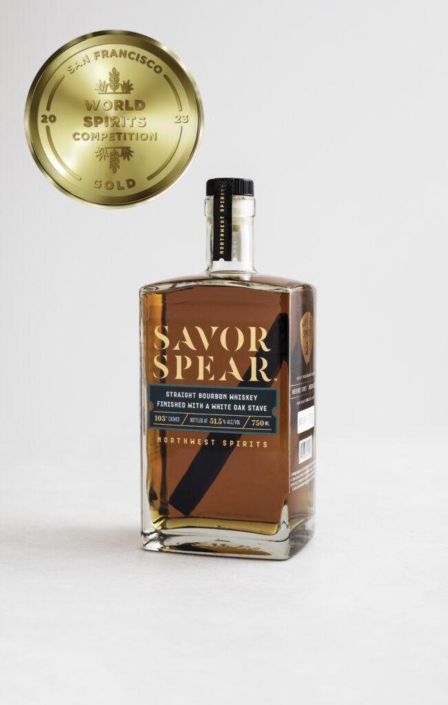 Image of a bottle of Savor Spear Bourbon Whiskey sitting on a clean white backdrop. The whiskey has an oak spear inside. There is a gold medal icon to the left of the bottle indicating the award that was won. 