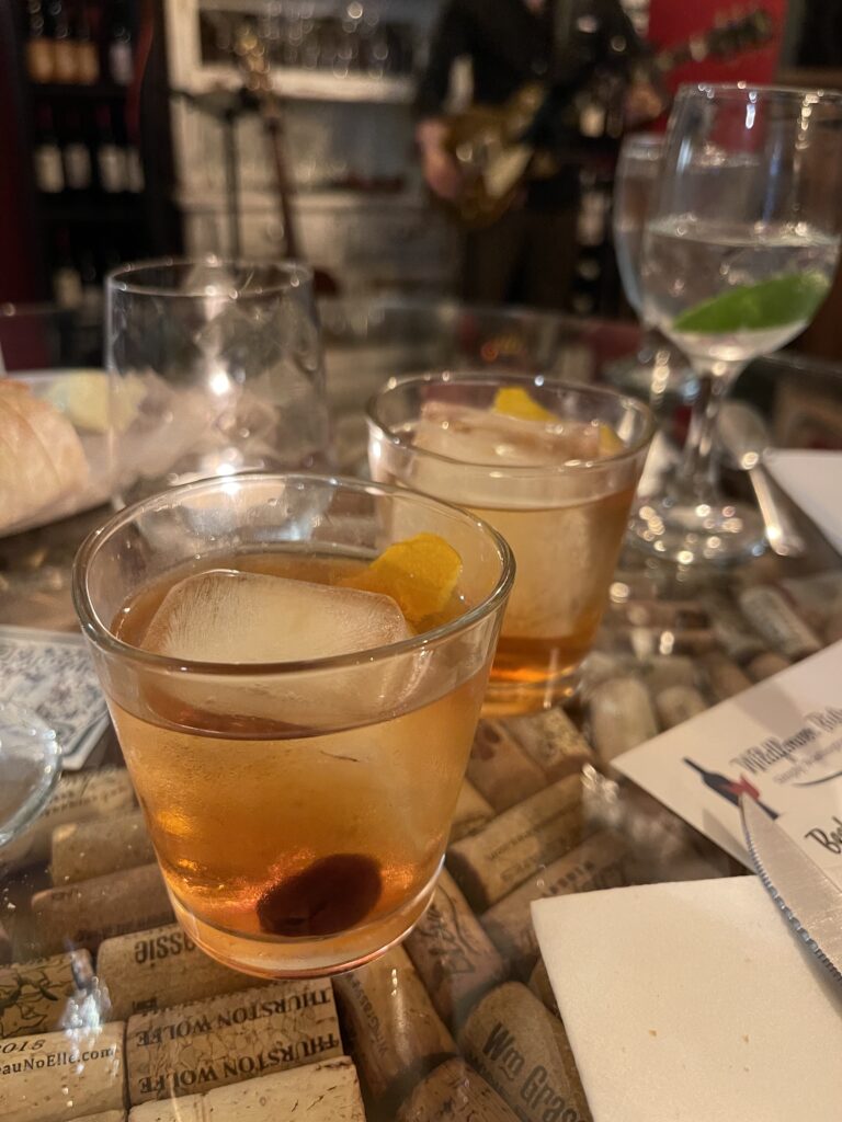 Photo of two manhattan cocktails on a glass table. Both manhattans have a large square block of ice, a bourbon cherry, and an orange peel.