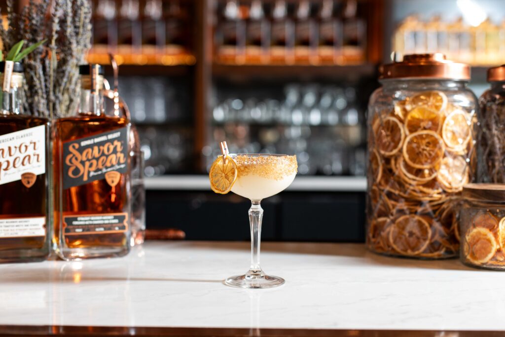 Lemon Confection cocktail in a coupe glass with a sugared rim and dried lemon garnish, surrounded by bottles of Savor Spear® and jars of citrus slices.