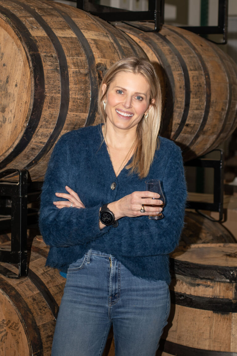 Merrisa Claridge, owner of Northwest Spirits, stands in front of a few wooden barrels. She's holding a glass of whiskey with her arms crossed, smiling.