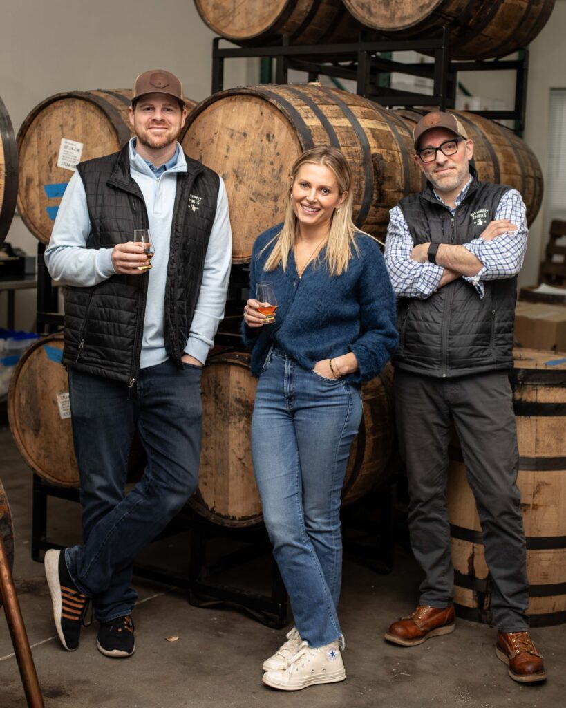 Merrisa Claridge, owner of Northwest Spirits, stands in front of a few wooden barrels beside her business partners, Nick and Jason. They are each holding a glass of whiskey, smiling.