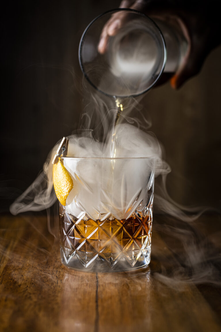 An impressive photo of a glass of whiskey being poured by hand. There is smoke billowing out from the decanter into the glass. The glass is garnished with an orange peel.