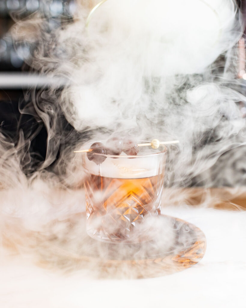 Smoked Black Manhattan cocktail in a crystal glass, garnished with cocktail cherries and surrounded by swirling aromatic smoke.