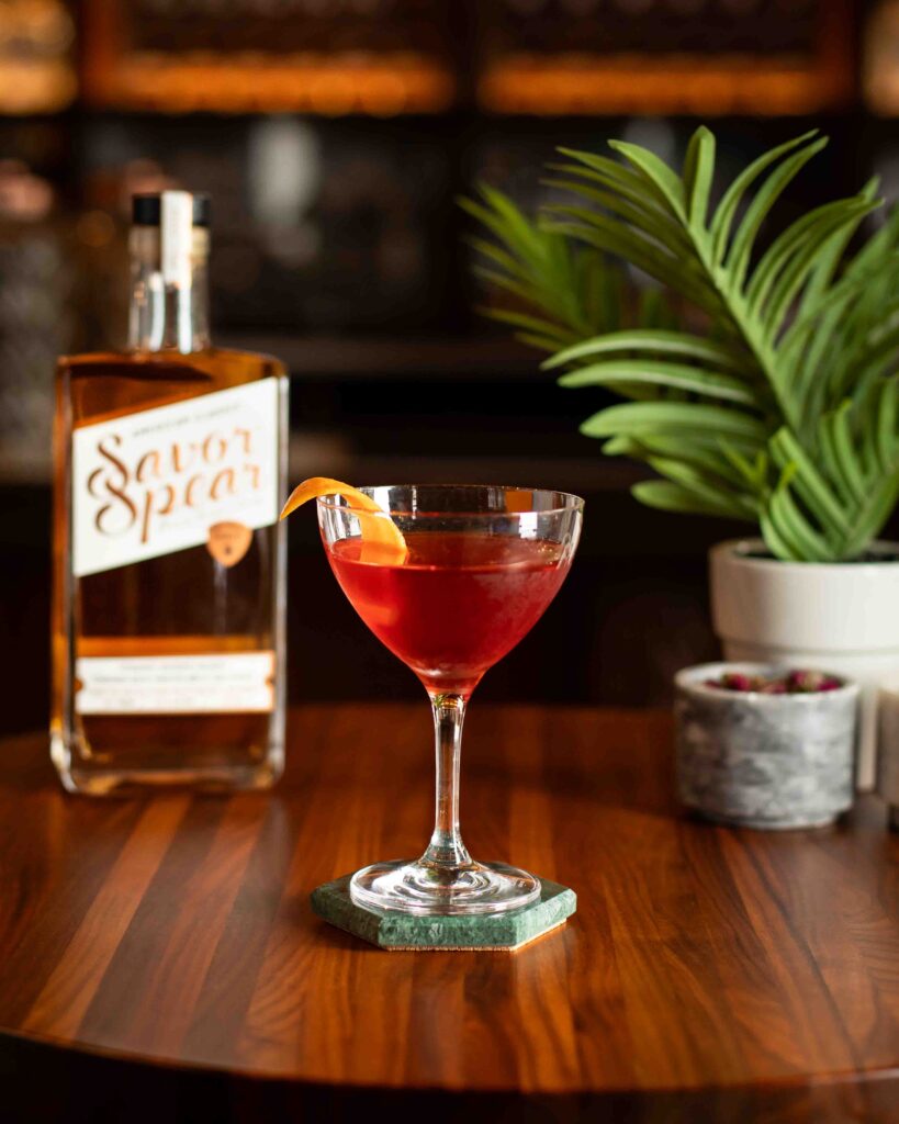 Barrel-aged Boulevardier cocktail in a coupe glass with orange twist garnish, served beside a bottle of Savor Spear® Bourbon.
