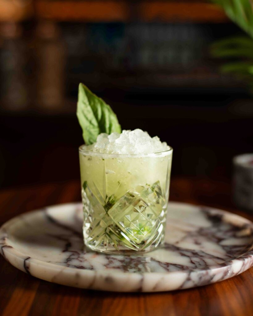 Basil Smash cocktail served over crushed ice in a crystal rocks glass, garnished with a fresh basil leaf and muddled herbs.