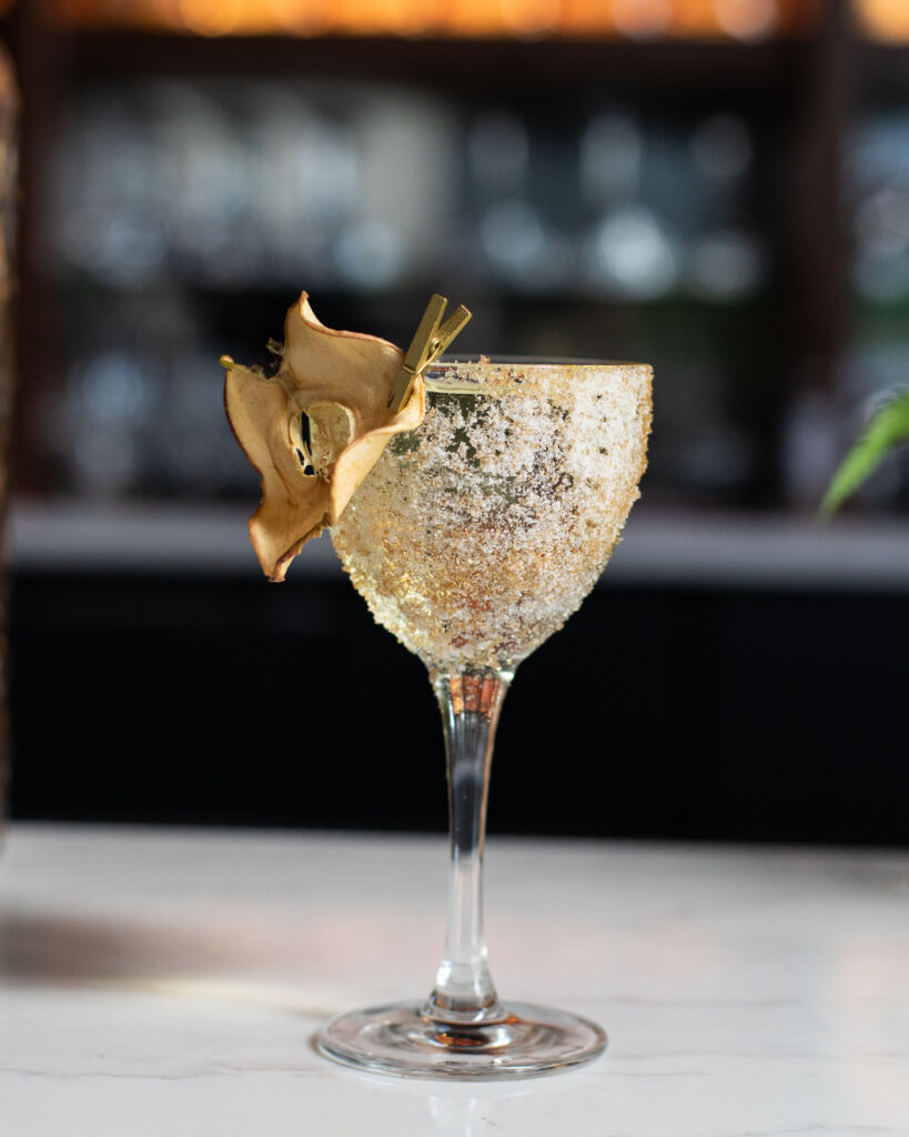 An elegant coupe glass coated in a sugar crust, garnished with a dried pear slice clipped to the rim.