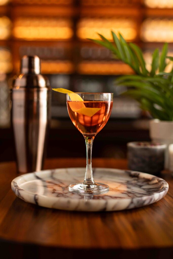Fast Penny Martinez cocktail in a coupe glass with lemon twist, set on a marble tray with a bar shaker in the background.