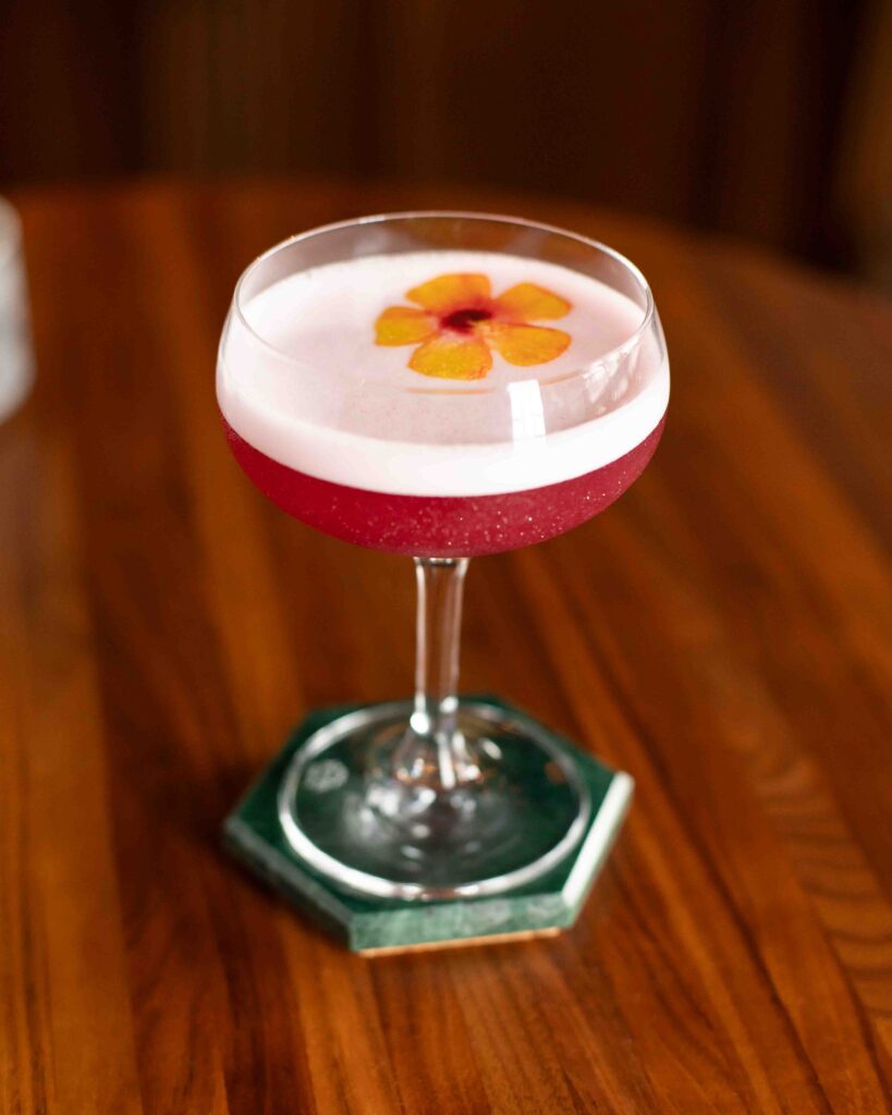 Hibiscus Sparkler cocktail with a frothy top and floral garnish, served in a coupe glass with a shimmering magenta hue.