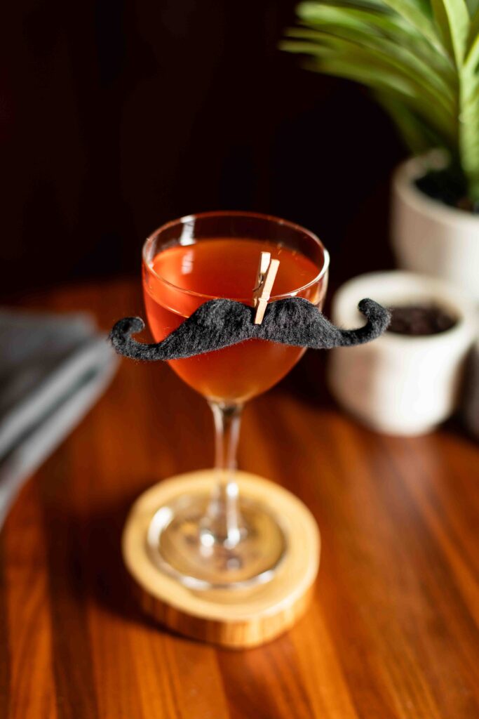 Amber bourbon cocktail in a coupe glass, garnished with a black felt mustache clipped to the rim.