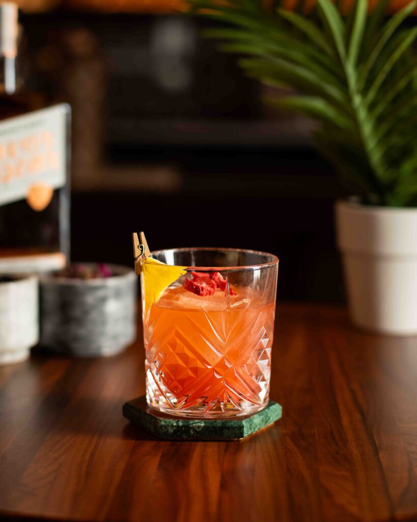 Strawberry and peach bourbon cocktail in a crystal glass, garnished with citrus peel and freeze-dried strawberry.