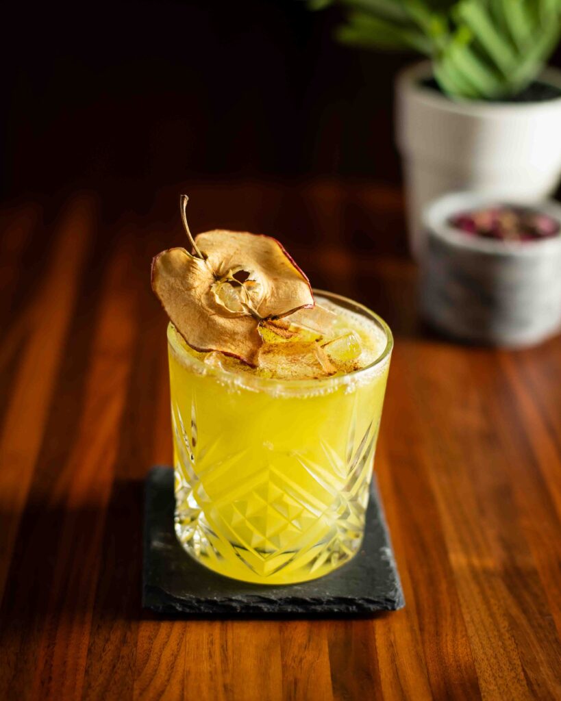 Bright green apple cocktail served over ice in a crystal glass, garnished with a dried apple slice.