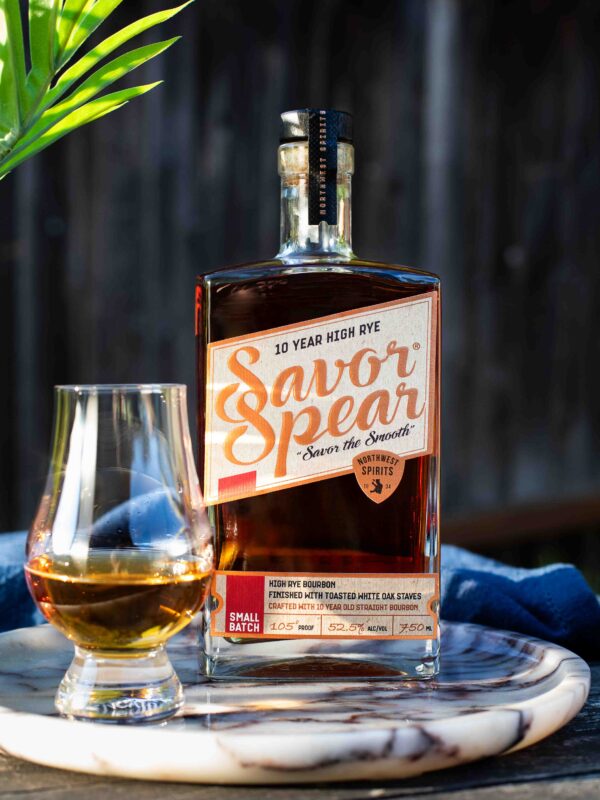 A bottle of Savor Spear® 10 Year High Rye Bourbon sits on a marble tray beside a poured Glencairn glass of bourbon. The label highlights its 105 proof, small batch production, and finish with toasted white oak staves. Sunlight catches the amber liquid, creating a warm, inviting glow, with a palm frond casting a soft shadow in the background.