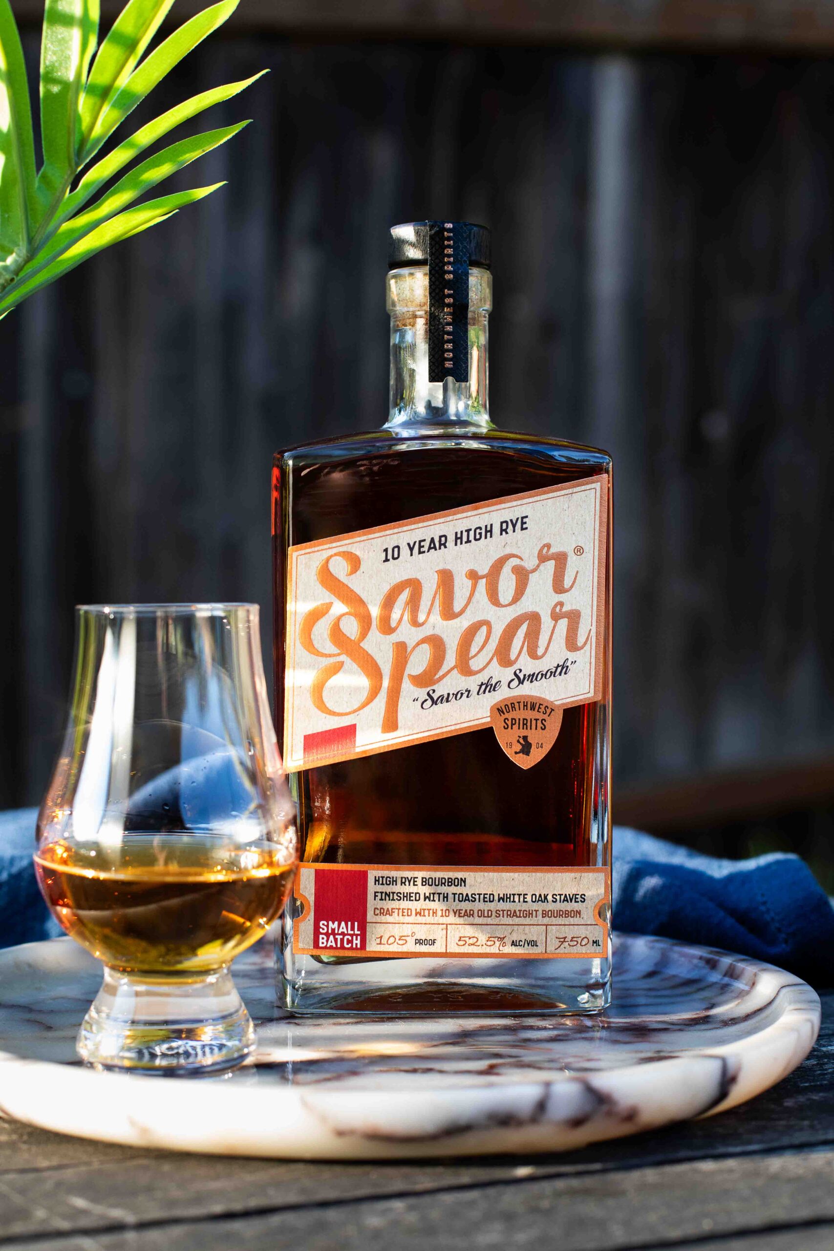 A bottle of Savor Spear® 10 Year High Rye Bourbon sits on a marble tray beside a poured Glencairn glass of bourbon. The label highlights its 105 proof, small batch production, and finish with toasted white oak staves. Sunlight catches the amber liquid, creating a warm, inviting glow, with a palm frond casting a soft shadow in the background.